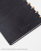A Warehouse Item - Discbound Heirloom Journal (Black/Brass) features full grain black leather with brass rings, displaying natural blemishes and irregular grain. Text at the bottom reads: Example of cosmetic blemish or irregular leather grain.