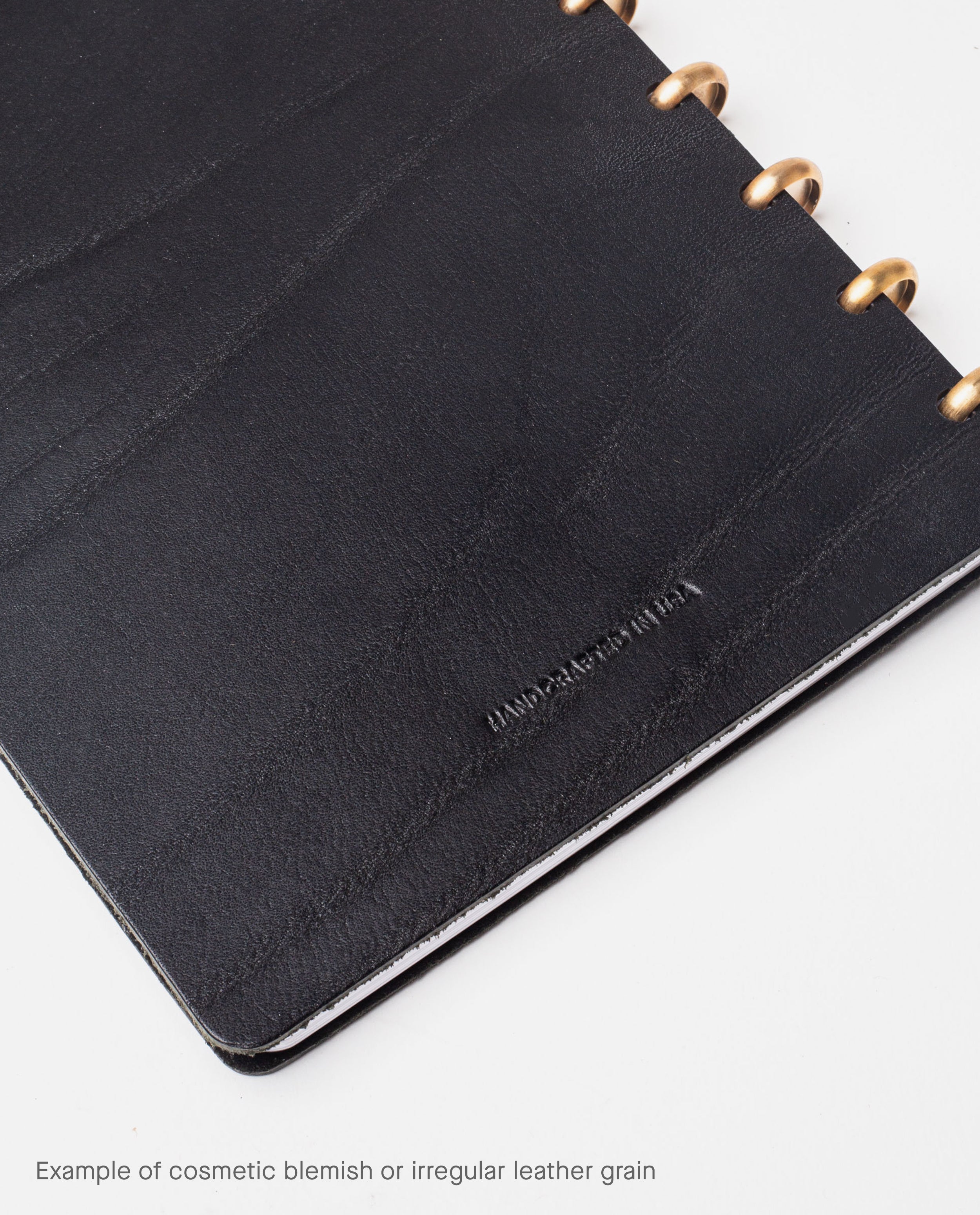 A Warehouse Item - Discbound Heirloom Journal (Black/Brass) features full grain black leather with brass rings, displaying natural blemishes and irregular grain. Text at the bottom reads: Example of cosmetic blemish or irregular leather grain.