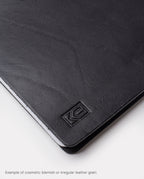 Close-up of the black full grain leather surface on the Warehouse Item - Discbound Heirloom Journal (Black/Brass), showing natural grain variations and a subtle embossed logo in the corner. Text below reads, Example of cosmetic blemish or irregular leather grain—perfect for our refillable discbound journal warehouse sale.