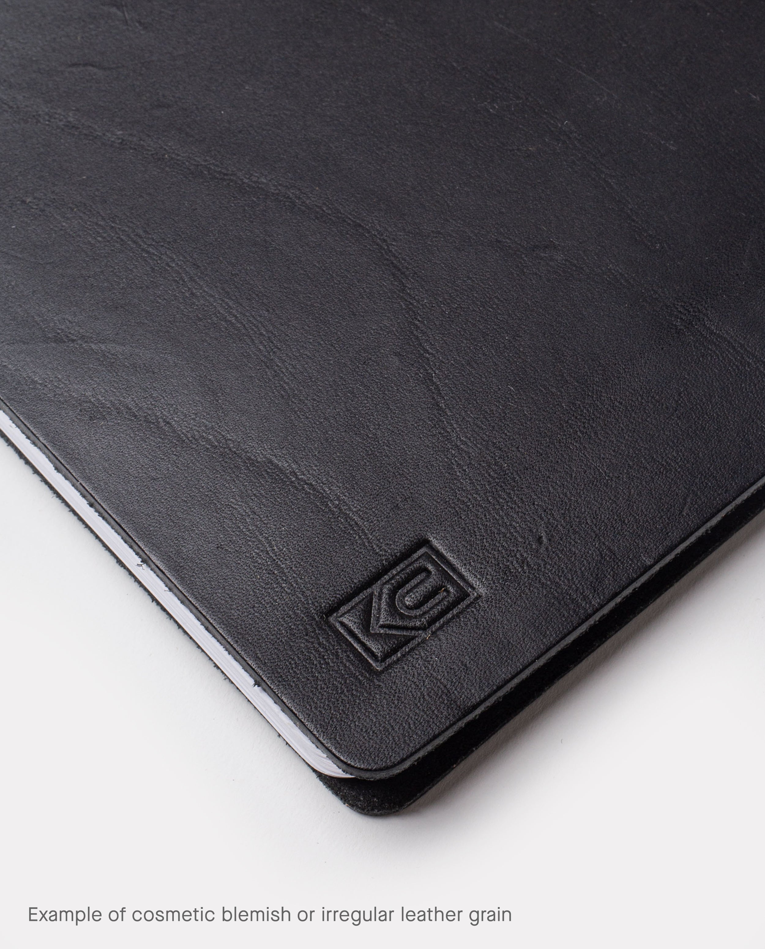 Close-up of the black full grain leather surface on the Warehouse Item - Discbound Heirloom Journal (Black/Brass), showing natural grain variations and a subtle embossed logo in the corner. Text below reads, Example of cosmetic blemish or irregular leather grain—perfect for our refillable discbound journal warehouse sale.