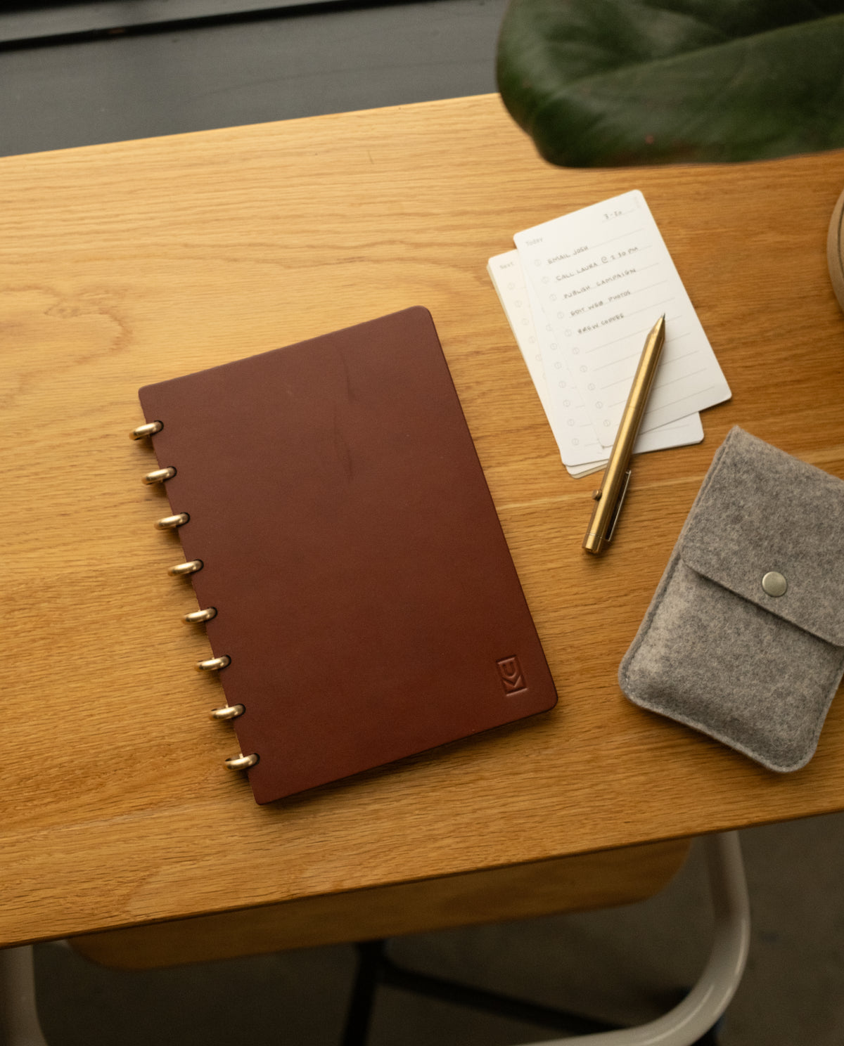 A Discbound Heirloom Journal in brown leather (6" x 8.5"), a gold pen, dot grid note cards with to-do list, and a gray felt pouch are arranged on a wooden desk beside a green plant.