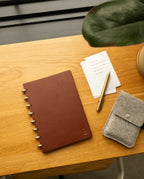 Here is a revised version of your sentence using the given product data: The Discbound Heirloom Journal Bundle, featuring brown leather in a 6" x 8.5" size with three refills, sits on a wooden desk next to a gold pen, lined note cards, a gray felt pouch, and a potted plant.