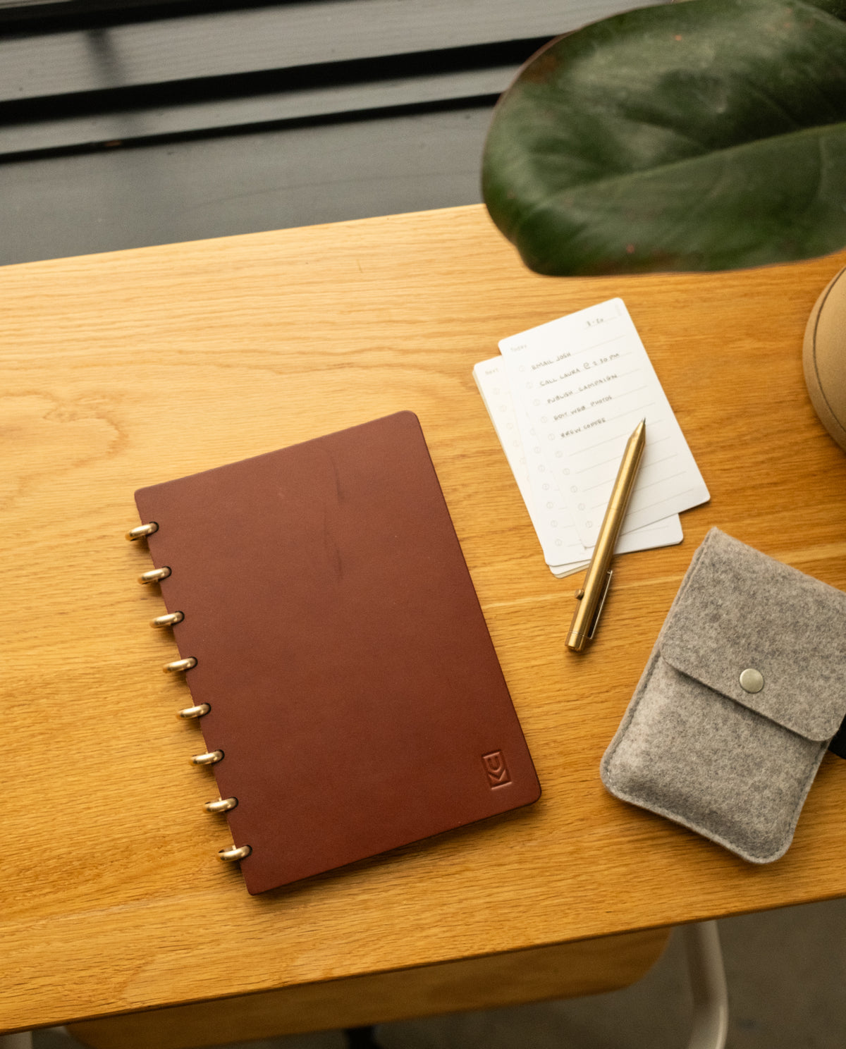 Here is a revised version of your sentence using the given product data: The Discbound Heirloom Journal Bundle, featuring brown leather in a 6" x 8.5" size with three refills, sits on a wooden desk next to a gold pen, lined note cards, a gray felt pouch, and a potted plant.