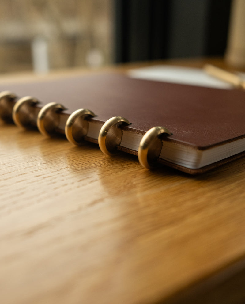 A close-up of the Discbound Heirloom Journal Bundle (Brown Leather 6" x 8.5" + 3 Refills) on a wooden table, featuring its gold rings, detailed cover edge, and lined refill pages inside to showcase the refillable discbound system.