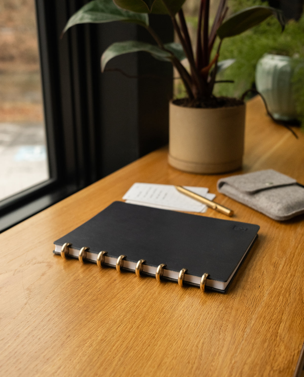 A Discbound Heirloom Journal (Black Leather - Standard 6" x 8.5"), a pen, and a small pouch sit on a wooden desk near a window, softly illuminated by sunlight with a potted plant in the background.