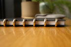 A close-up side view of the Discbound Heirloom Journal (Black Leather - Standard 6" x 8.5") with gold disc rings resting on a wooden table, blurred objects in the background.