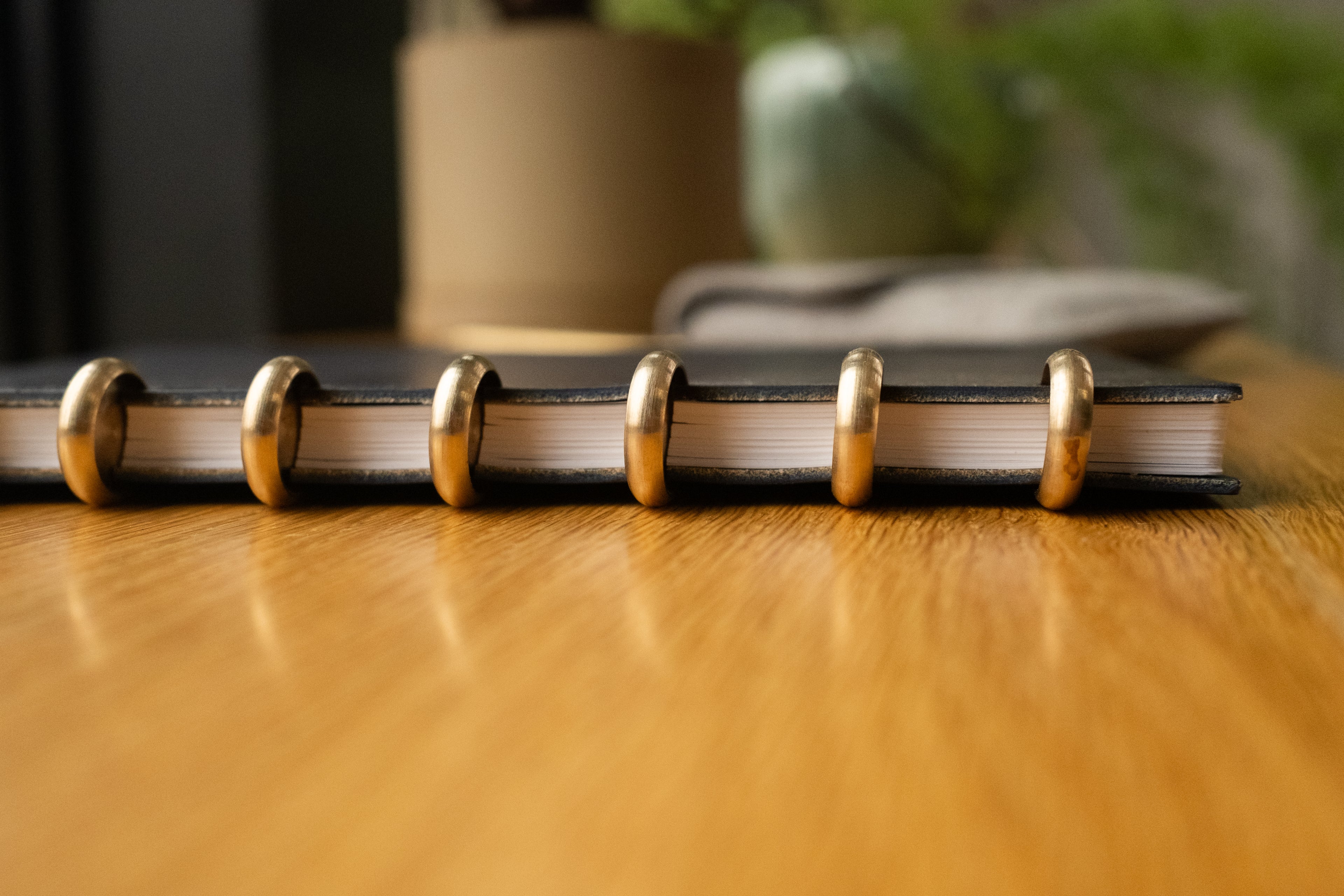 A close-up side view of the Discbound Heirloom Journal (Black Leather - Standard 6" x 8.5") with gold disc rings resting on a wooden table, blurred objects in the background.