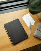 A Discbound Heirloom Journal Bundle (Black Leather - Standard 6" x 8.5" + 3 Refills) sits on a wooden desk near a window, accompanied by a silver pen, lined note cards, part of a potted plant, and a gray felt pouch.
