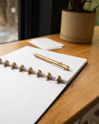 A Discbound Heirloom Journal (Black Leather - Standard 6" x 8.5") and a gold pen rest on a wooden desk by a window, with a potted plant and small stack of cards in the background.