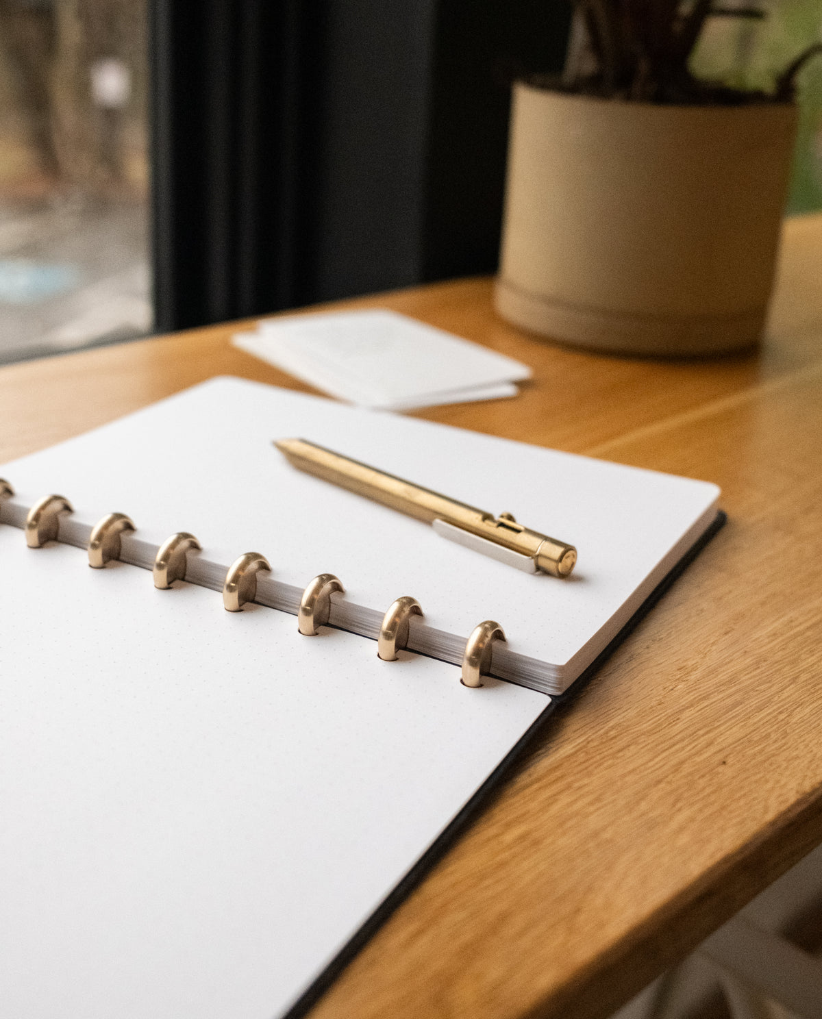 A Discbound Heirloom Journal (Black Leather - Standard 6" x 8.5") and a gold pen rest on a wooden desk by a window, with a potted plant and small stack of cards in the background.