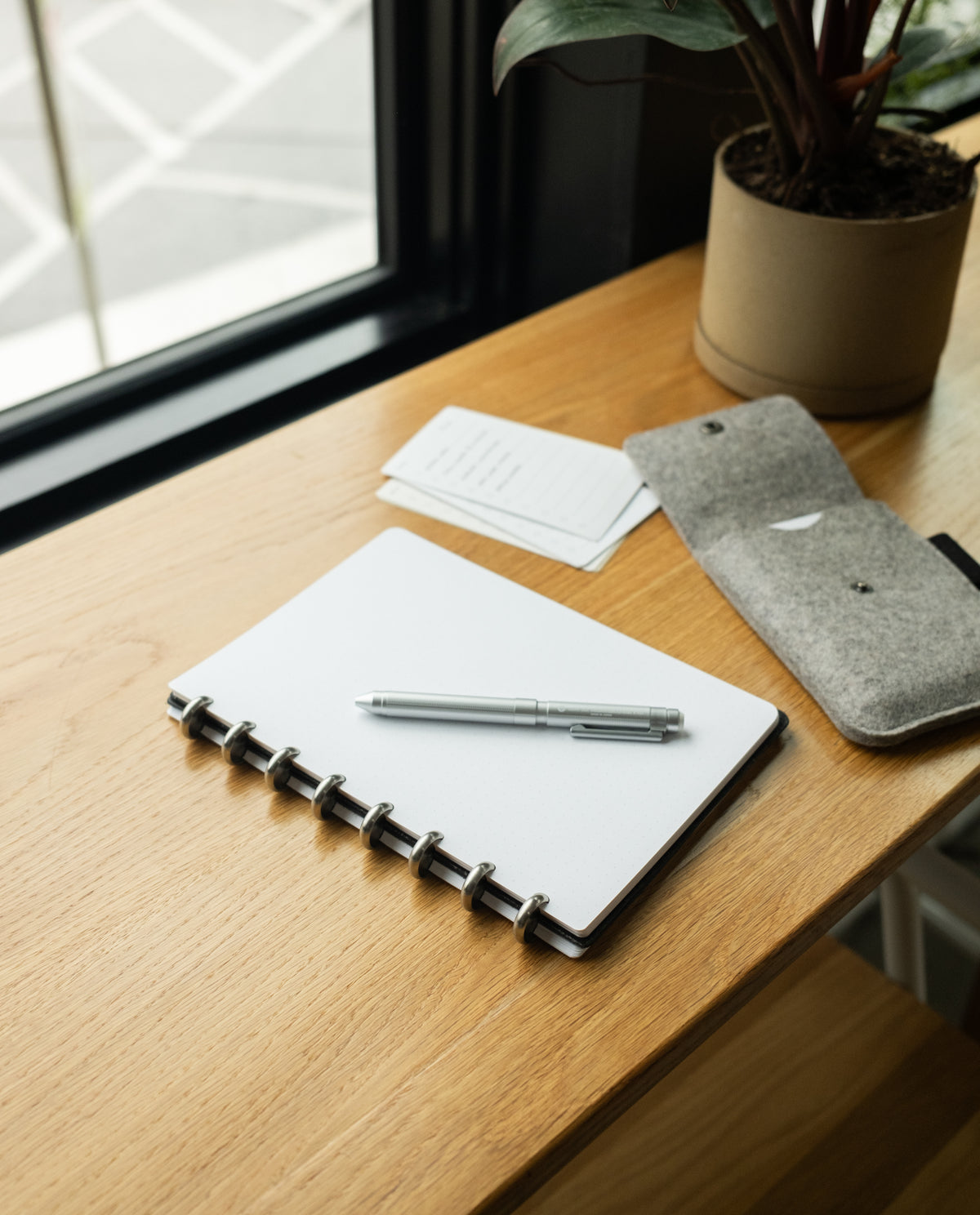 A silver pen rests on a black leather Discbound Heirloom Journal Bundle (Standard 6" x 8.5" + 3 Refills) on a wooden desk near a window, with a potted plant, felt pen case, and note cards in sunlight.