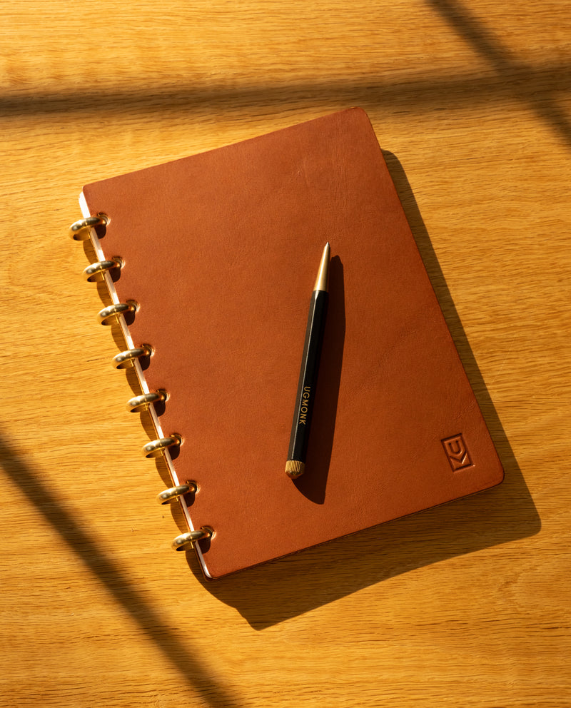 The Discbound Heirloom Journal in tan leather (Standard 6" x 8.5") rests on a wooden surface with diagonal shadows, gold rings glinting and a black and gold pen placed neatly on top.