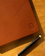 A Discbound Heirloom Journal (Tan Leather - Standard 6" x 8.5") with an embossed logo sits on a wooden surface next to a black pen with gold accents, bathed in sunlight and warm shadows.