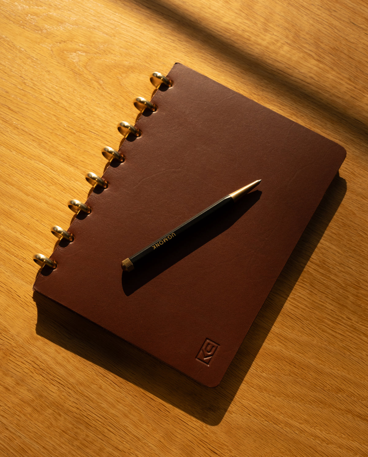 A black and gold pen rests on a Discbound Heirloom Journal in brown full-grain leather with gold rings, placed on a wooden surface in warm sunlight.