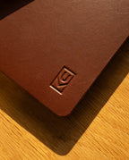 A close-up of the Discbound Heirloom Journal in brown full-grain leather, featuring an embossed geometric logo on the cover, rests on a wooden surface bathed in warm sunlight. Inside, this standard 6" x 8.5" refillable journal includes premium dot grid paper for versatile note-taking.