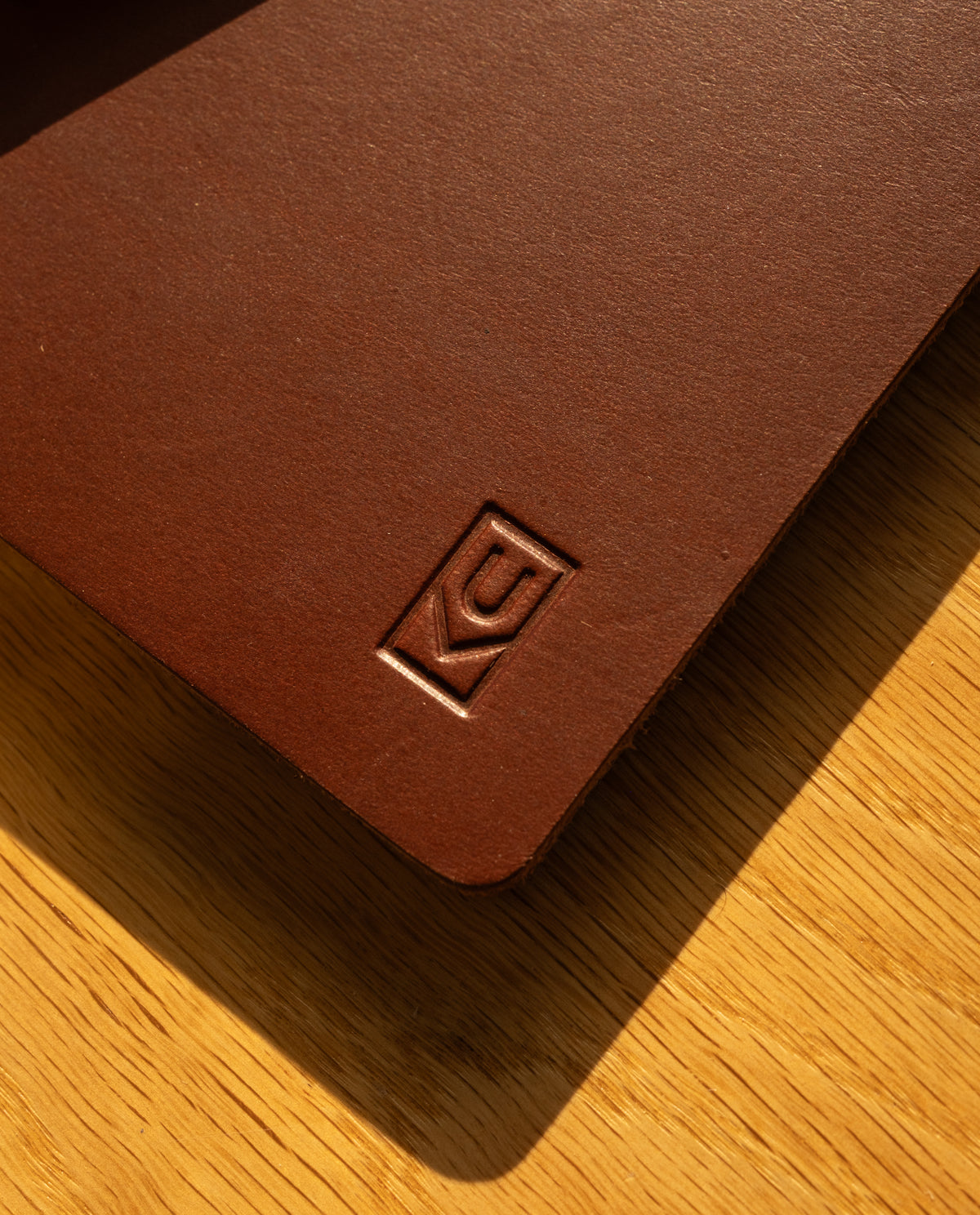 A close-up of the Discbound Heirloom Journal in brown full-grain leather, featuring an embossed geometric logo on the cover, rests on a wooden surface bathed in warm sunlight. Inside, this standard 6" x 8.5" refillable journal includes premium dot grid paper for versatile note-taking.