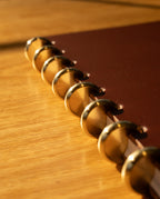 Close-up of a Discbound Heirloom Journal (Brown Leather - Standard 6" x 8.5") featuring full-grain brown leather and gold-colored disc binding rings, resting on a wooden surface bathed in warm sunlight.