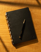 A Discbound Heirloom Journal (Black Leather - Standard 6" x 8.5") and a matching black pen rest on a wooden surface, bathed in warm sunlight and soft shadows.