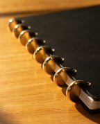 A close-up of the Discbound Heirloom Journal (Black Leather - Standard 6" x 8.5") with gold disc binding rings, resting on a wooden surface in warm sunlight, highlighting the edge and elegant, refillable rings.
