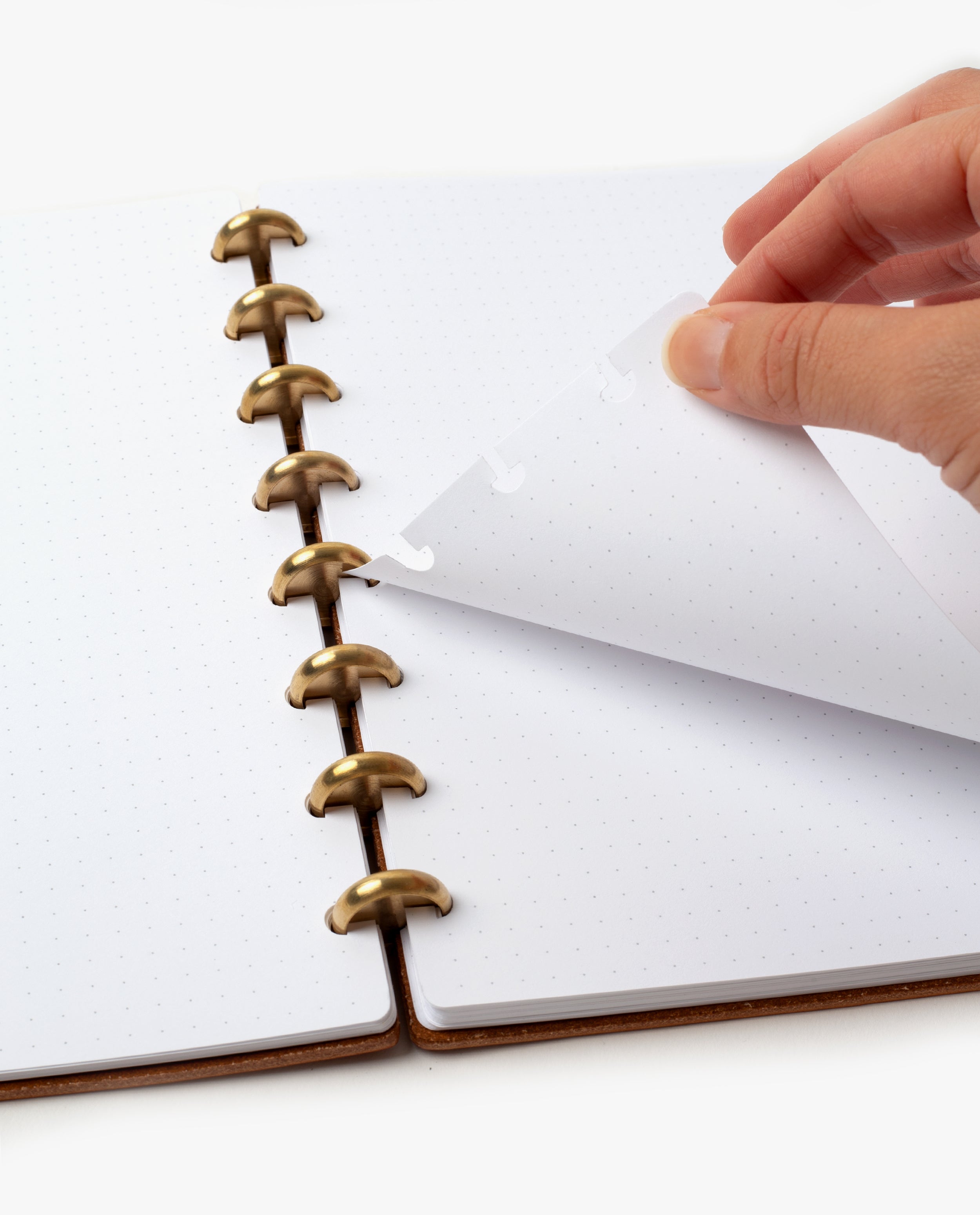A hand lifts a dotted page from the Discbound Heirloom Journal (Tan Leather - Standard 6" x 8.5") with gold disc binding, featuring removable and repositionable dot grid paper against a white background.