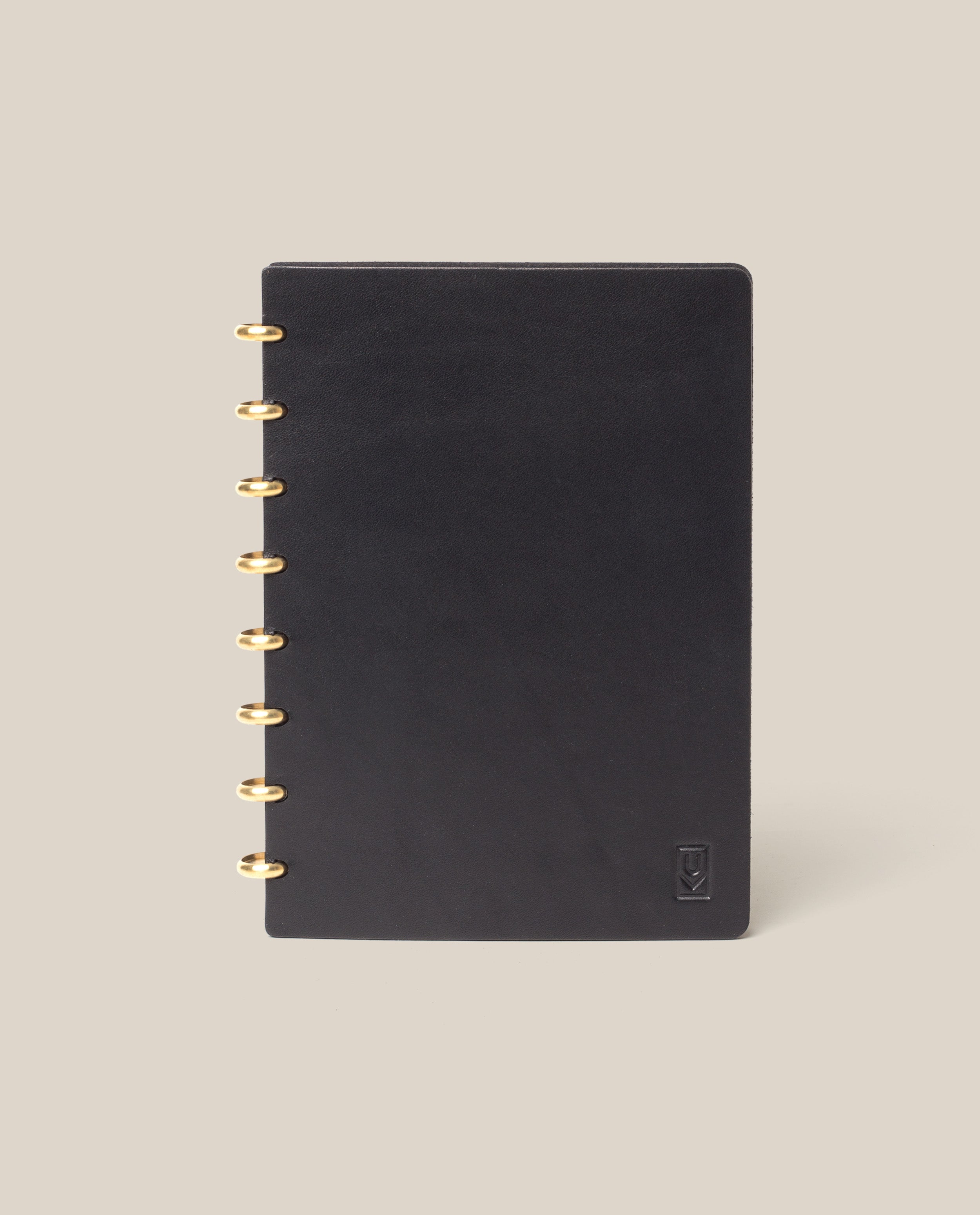 The Discbound Heirloom Journal (Black Leather - Standard 6" x 8.5") with gold rings is centered on a beige background, featuring a small embossed logo in the lower right corner of its black leather cover.