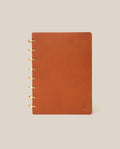 The Discbound Heirloom Journal (Tan Leather - Standard 6" x 8.5") stands upright on a plain beige background, featuring gold ring binding on the left and a small embossed logo in the bottom right corner of its refillable leather cover.