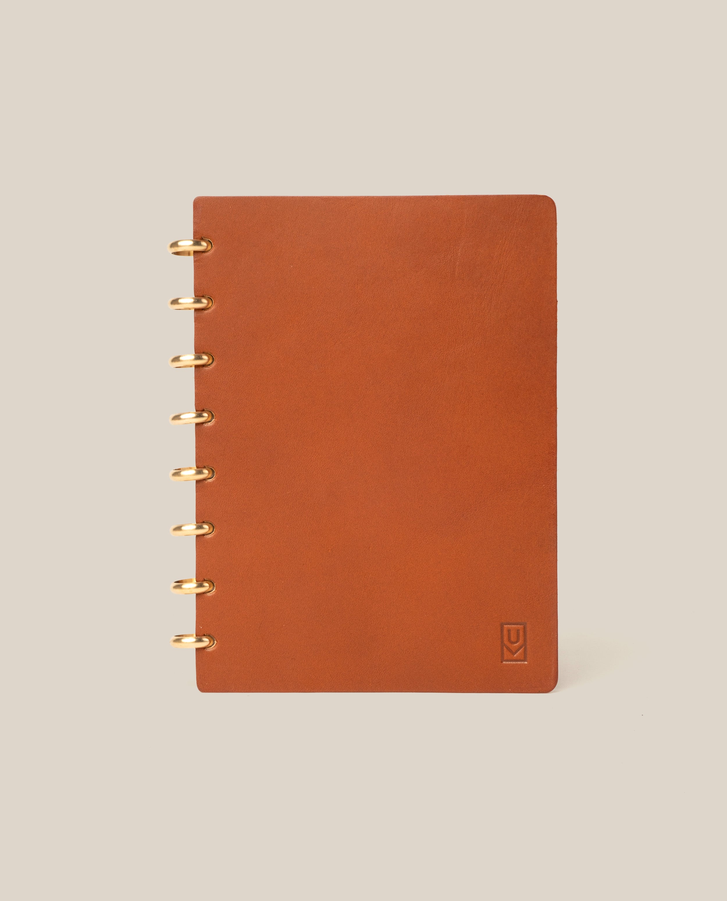 The Discbound Heirloom Journal (Tan Leather - Standard 6" x 8.5") stands upright on a plain beige background, featuring gold ring binding on the left and a small embossed logo in the bottom right corner of its refillable leather cover.