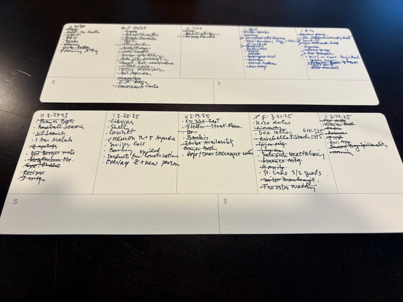 Two handwritten task lists on white notecards are laid side by side on a dark surface. Each card is divided into columns with various notes, to-dos, and checklists written in black ink.
