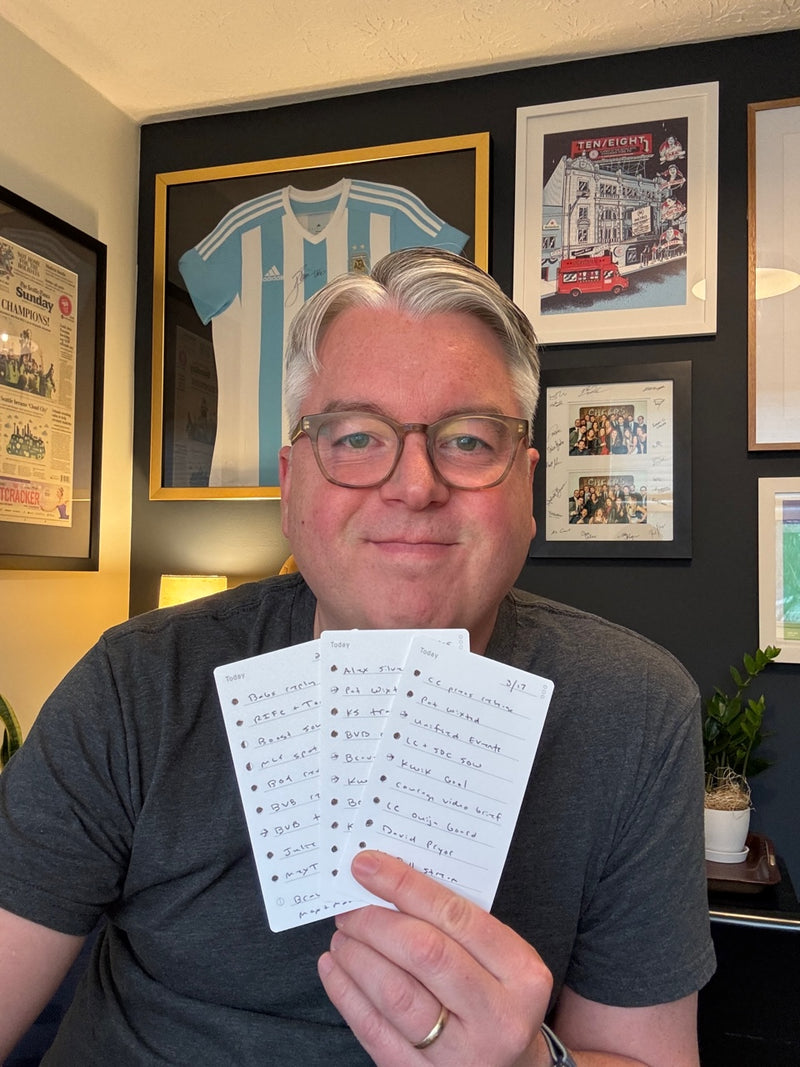 A man with gray hair and glasses smiles while holding up three handwritten note cards. Behind him are framed sports memorabilia, including a blue and white striped jersey.