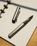A graphite LAMY AL-star Rollerball with its cap off rests on an open, dotted notebook, demonstrating smooth writing in sunlight that casts shadows across the wooden surface.