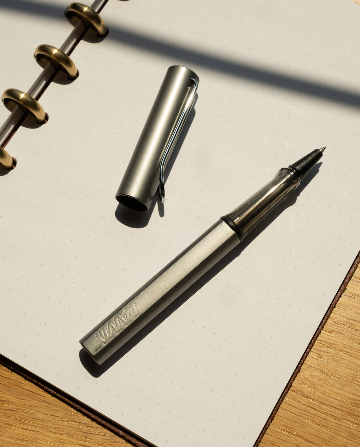 A graphite LAMY AL-star Rollerball with its cap off rests on an open, dotted notebook, demonstrating smooth writing in sunlight that casts shadows across the wooden surface.