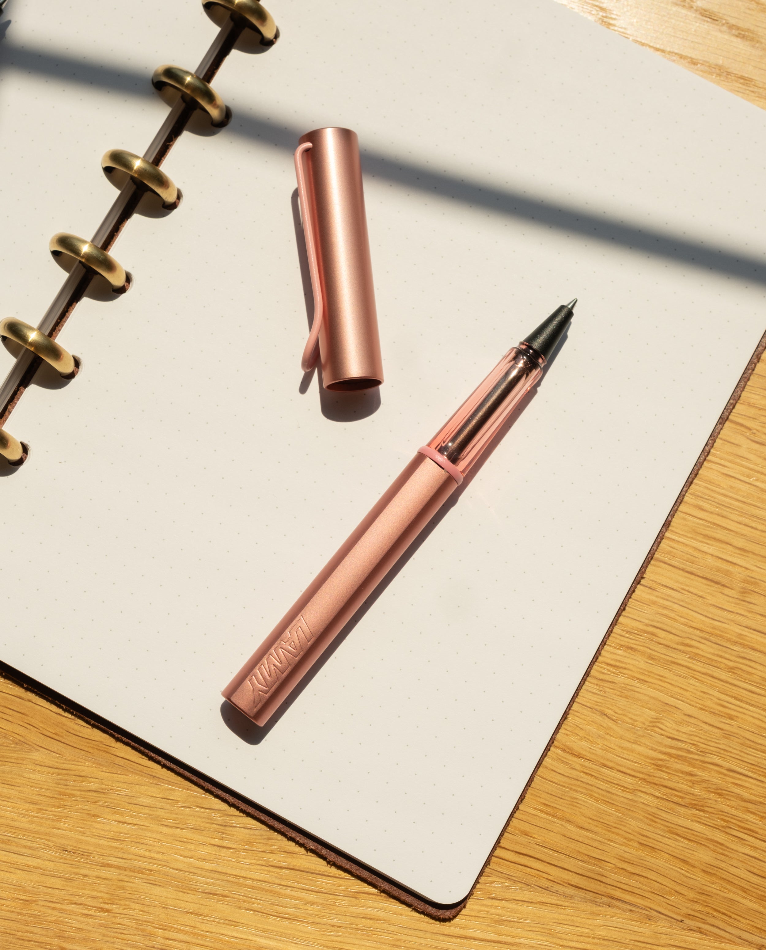 A LAMY AL-star Rollerball (Flamingo), cap off, rests on an open dotted spiral notebook atop a wooden surface, its anodized aluminum body gleaming in natural sunlight.