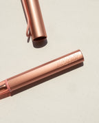 A close-up of the LAMY AL-star Rollerball (Flamingo) with its cap removed shows its anodized aluminum body on a light dotted surface, and "LAMY" embossed on the barrel.