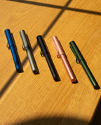 A LAMY AL-star Rollerball (Graphite) is displayed on a wooden surface with sunlight and shadows, highlighting its smooth writing performance and refillable rollerball design.
