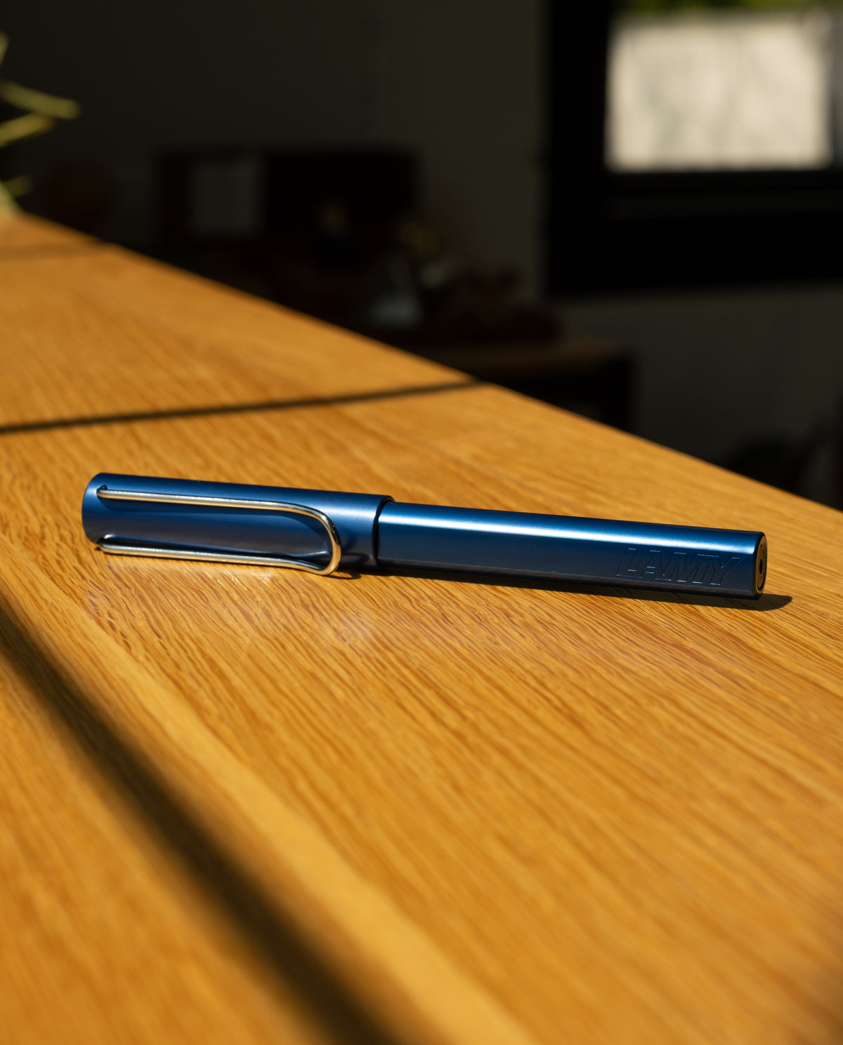 A LAMY AL-star Rollerball (Oceanblue) sits on a wooden surface in natural sunlight, its sleek design highlighted against a softly blurred background with a window and indoor decor.