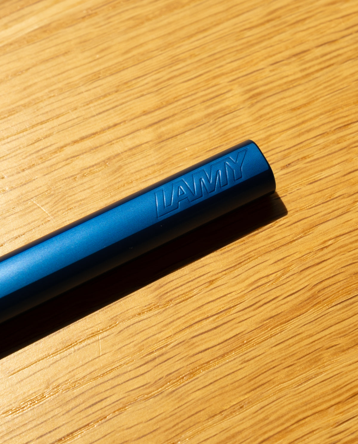 A close-up of a blue LAMY AL-star Rollerball (Oceanblue), the refillable pen known for smooth writing, embossed with the brand name and resting diagonally on a wooden surface in bright sunlight.