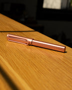The LAMY AL-star Rollerball (Flamingo), featuring a metallic pink anodized aluminum body, lies on a wooden surface in sunlight, casting gentle shadows and highlighting its modern design.