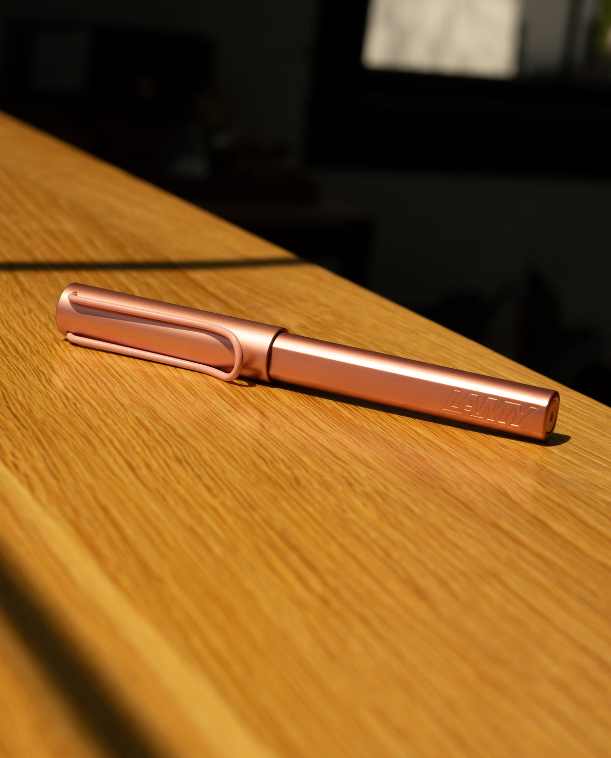The LAMY AL-star Rollerball (Flamingo), featuring a metallic pink anodized aluminum body, lies on a wooden surface in sunlight, casting gentle shadows and highlighting its modern design.