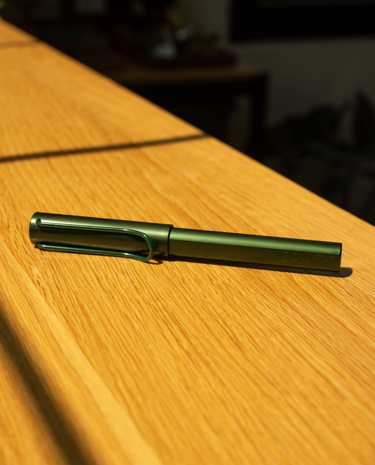 A LAMY AL-star Rollerball (Pine) rests on a sunlit wooden surface, casting a rightward shadow—a refillable rollerball pen recognized for its smooth writing experience.