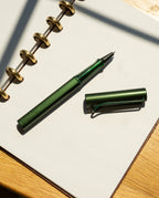 A pine green LAMY AL-star Rollerball with its cap off rests on an open dotted notebook with gold rings, placed on a sunlit wooden surface—perfect for smooth writing.