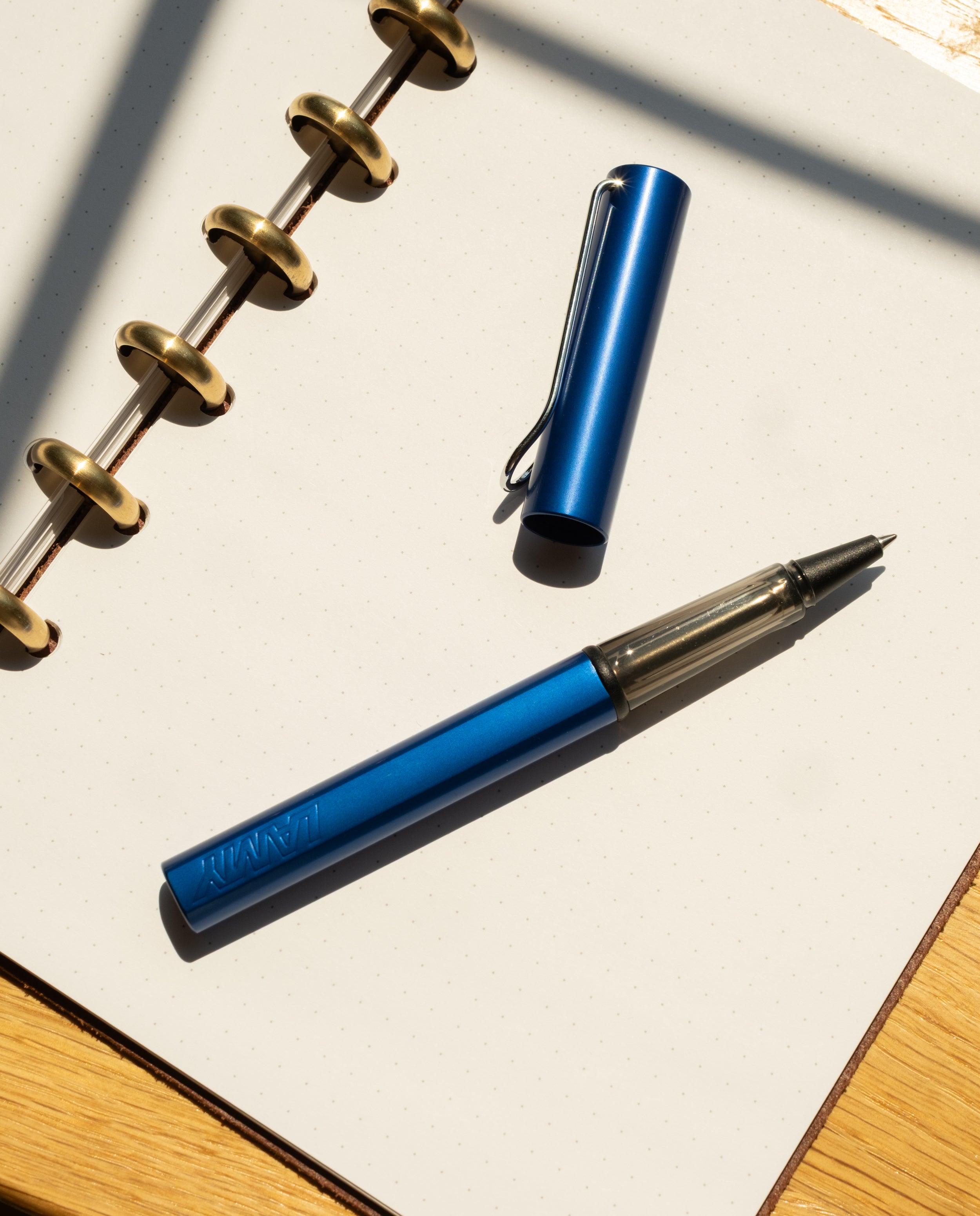 A LAMY AL-star Rollerball (Oceanblue) with its cap off rests on an open dotted notebook with gold rings, set on a wooden surface in bright sunlight, highlighting its smooth writing and refillable rollerball design.