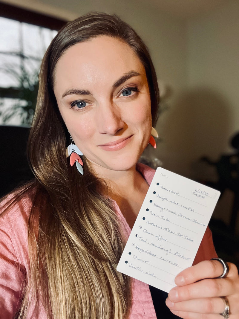 A woman with long brown hair, wearing a pink shirt and colorful earrings, smiles while holding a handwritten to-do list in her hand. The background is softly blurred.