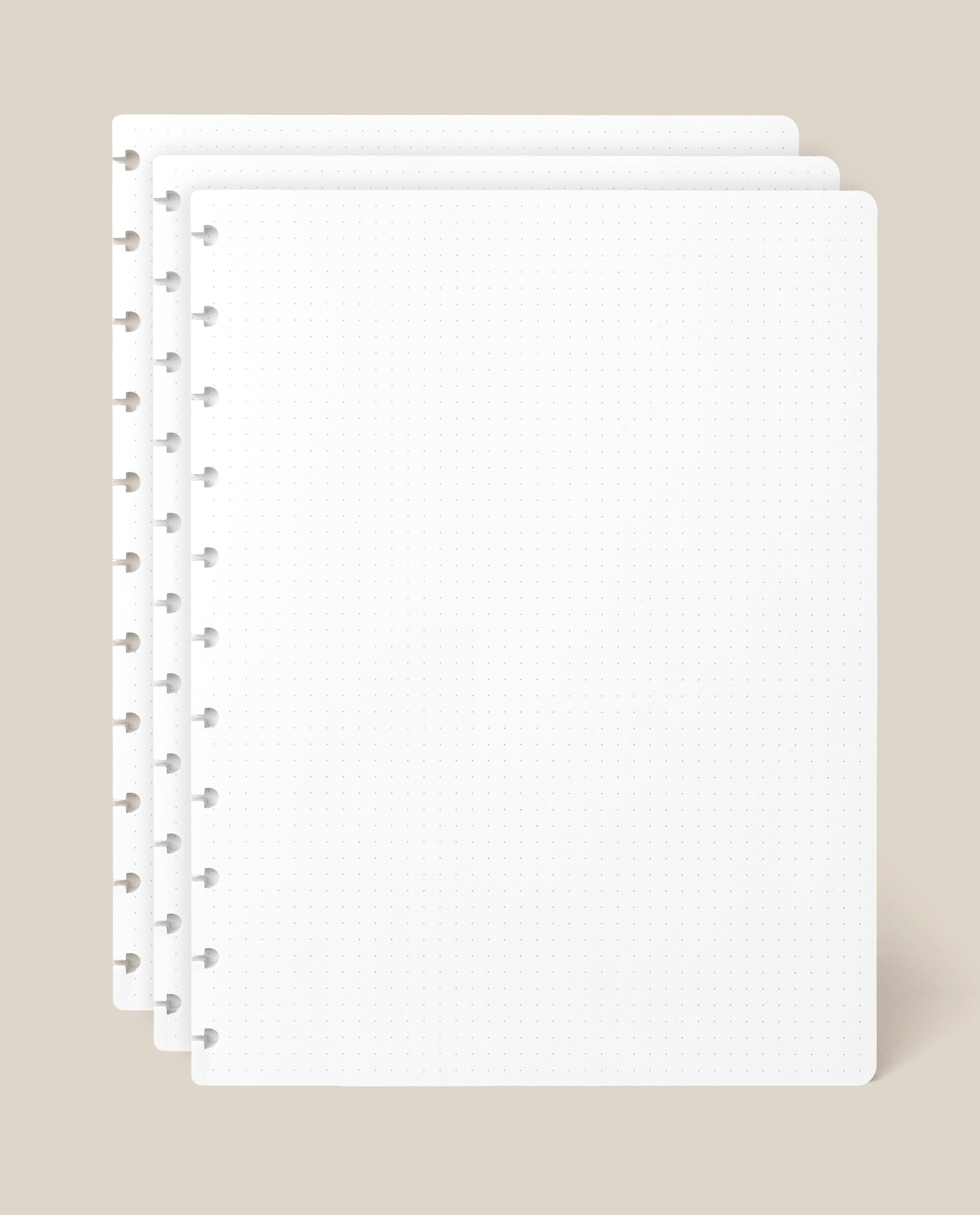 Three sheets of the Large Discbound Journal Refill - Dot Grid (8.5″ x 11″)—white and perforated—are stacked with offset edges on a beige background, making them ideal for expanding your discbound notebook.