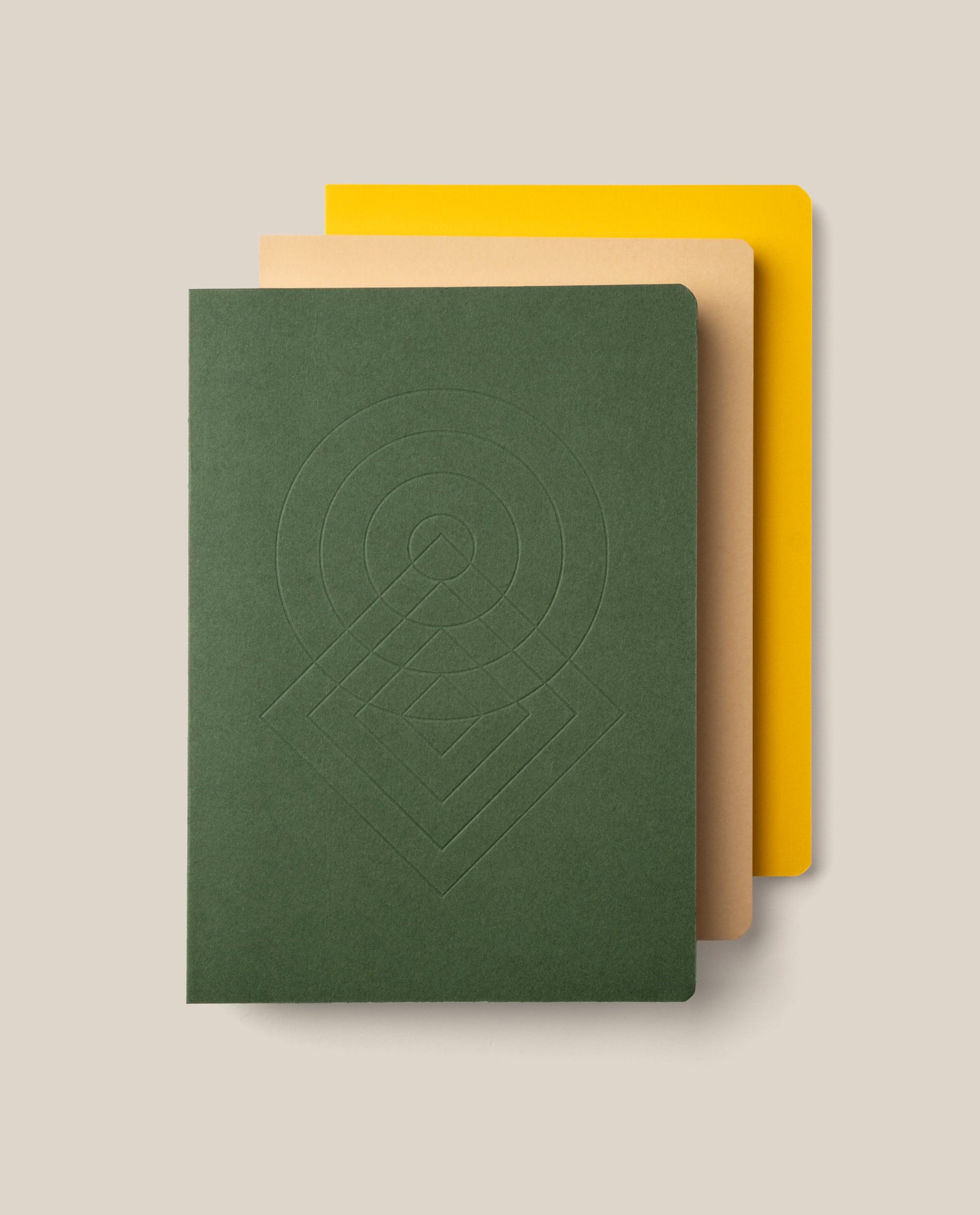 A set of three Layflat Notebook Bundle (Set of 3) notebooks are stacked on a beige background, featuring a dark green notebook with an embossed geometric pattern on top, a tan notebook in the middle, and a yellow notebook on the bottom. Each has rounded corners and durable Smyth sewn bindings that allow for effortless writing across every page.