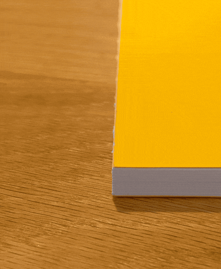 A hand taps the top corner of a yellow-covered Layflat Notebook Bundle (Set of 3) resting on a wooden table, making the notebook’s corner bounce slightly.