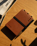 The Leather Discbound Analog + 3 Refills (Tan), featuring full-grain leather covers in a compact size with metal rings and secured by a black elastic band, sits alongside a black-and-gold pen on a wooden table bathed in warm sunlight.