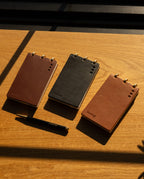 Three tan full-grain leather discbound notepads from the Leather Discbound Analog + 3 Refills set rest on a wooden surface, accompanied by a black pen and illuminated by sunlight casting gentle shadows across the scene.