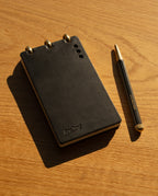 A Leather Discbound Analog + 3 Refills (Black), featuring full-grain leather covers and metal rings, rests on a wooden surface beside a black and gold pen. Sunlight casts shadows across the scene, highlighting the timeless elegance of this accessory.