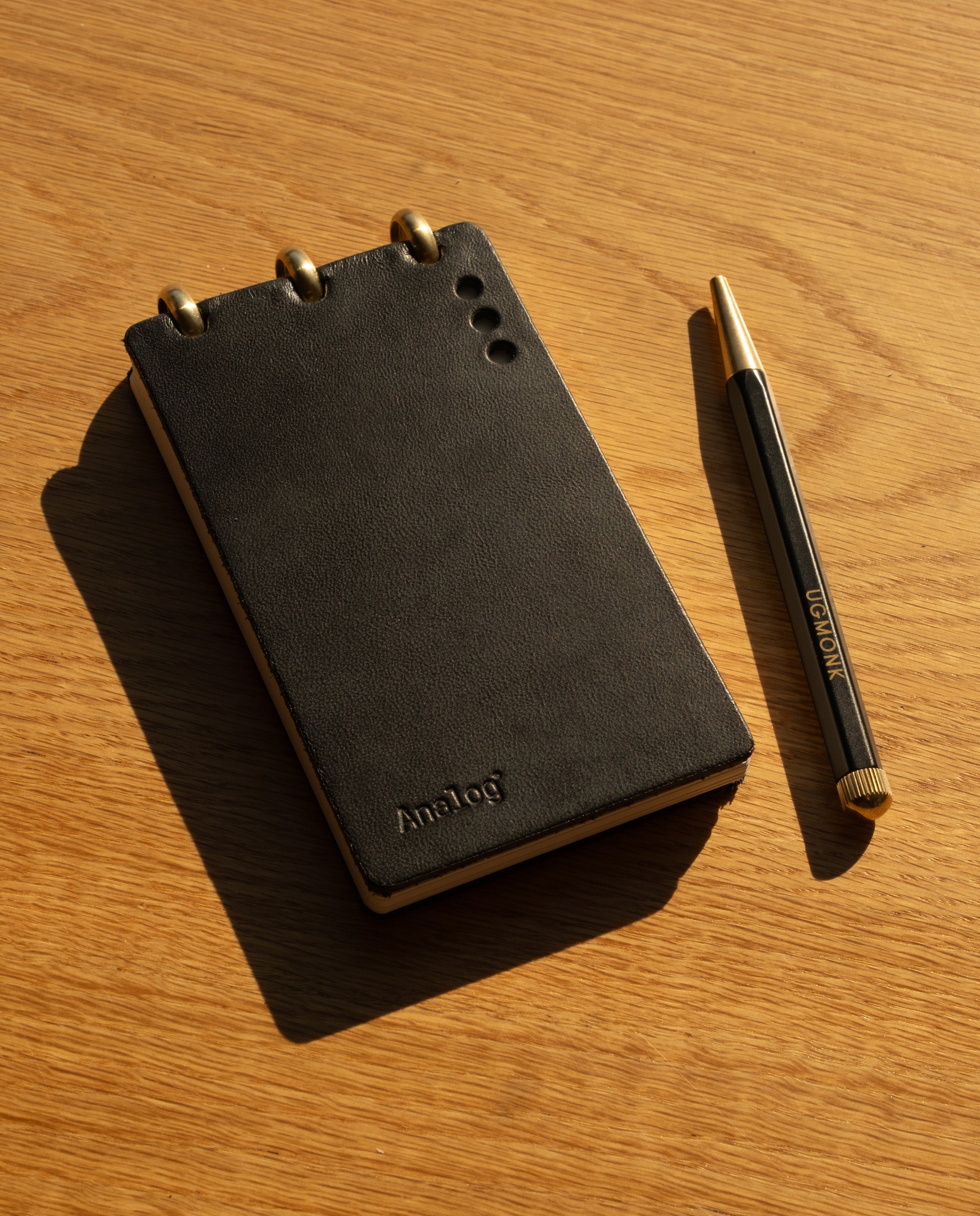 A Leather Discbound Analog + 3 Refills (Black), featuring full-grain leather covers and metal rings, rests on a wooden surface beside a black and gold pen. Sunlight casts shadows across the scene, highlighting the timeless elegance of this accessory.