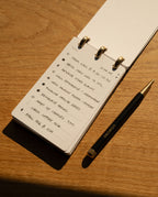 On a desk bathed in warm sunlight sits the Leather Discbound Analog + 3 Refills (Black), its full-grain leather covers lending a sophisticated touch to the daily to-do lists inside. Beside it rests a black and gold UCONIK pen.